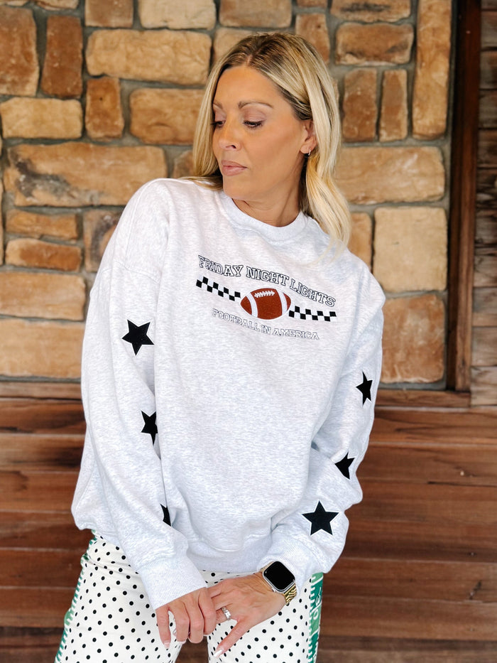 A woman wearing a light gray crewneck sweatshirt with black star prints and a football graphic that reads 'Tulsa Southwest Football Club'. She is standing in front of a rustic stone wall and wooden door.