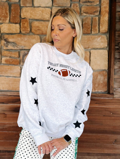 A woman wearing a light gray crewneck sweatshirt with black star prints and a football graphic that reads 'Tulsa Southwest Football Club'. She is standing in front of a rustic stone wall and wooden door.