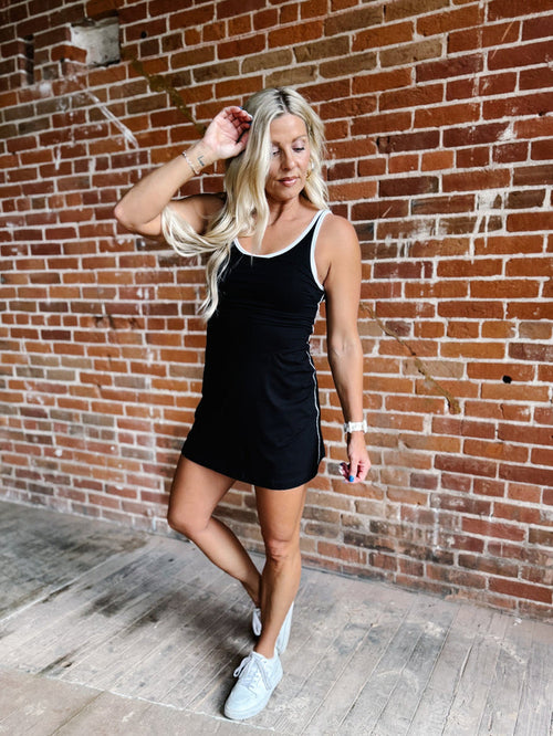 A woman wearing a black skortsie dress with a scoop neckline and contrast trim, stylishly posing indoors.