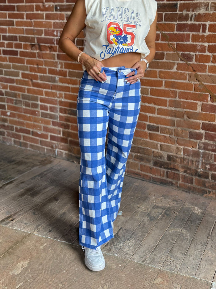 A person wearing blue and white gingham wide-leg pants, a cream sleeveless crop top with a colorful 'Kansas Jayhawks 65' graphic, and white sneakers, standing against a brick wall.