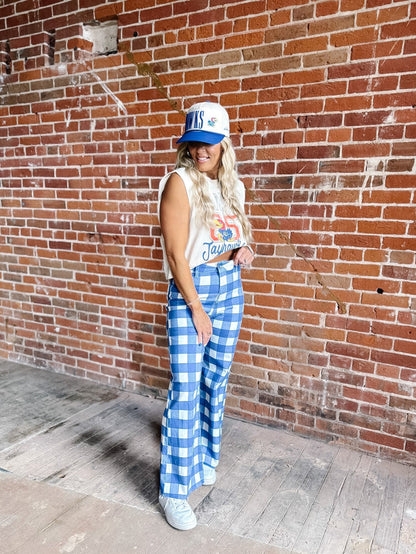 A woman stands against a red brick wall wearing a white and blue gingham checkered pair of wide-leg pants, a white graphic tank top, a blue and white baseball cap, and white sneakers.