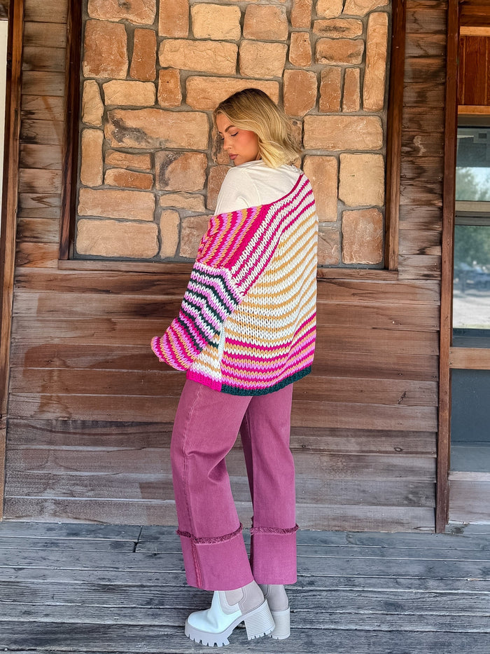 A woman stands on a wooden porch in front of a stone wall, wearing a chunky knit striped cardigan with bright pink, orange, yellow, white, and blue stripes, paired with dusty rose pants and white platform shoes.