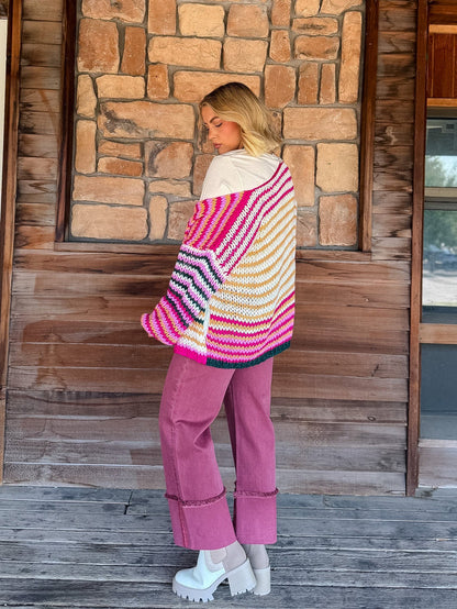 A woman stands on a wooden porch in front of a stone wall, wearing a chunky knit striped cardigan with bright pink, orange, yellow, white, and blue stripes, paired with dusty rose pants and white platform shoes.