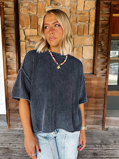 A woman with blonde hair wearing a Navy ribbed short-sleeve top and light blue jeans, standing on a wooden porch in front of a stone wall.
