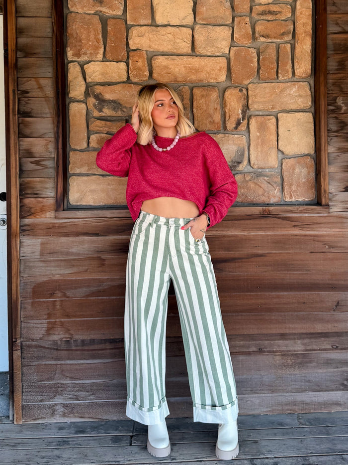 A woman stands on a wooden porch in front of a stone wall, wearing a bright pink cropped long-sleeve sweater, sage green and white vertically striped wide-leg jeans, and white boots.