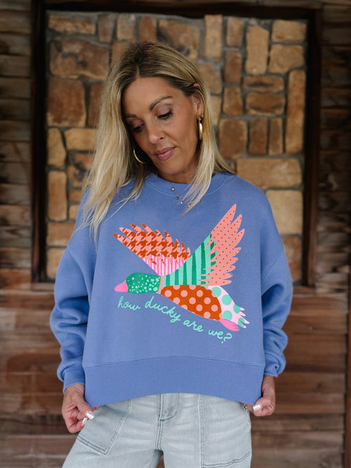 A woman wearing a blue crewneck sweatshirt with a colorful patchwork bird graphic and the text 'how ducky are we?' on the front, paired with light gray jeans, standing in front of a rustic stone wall.