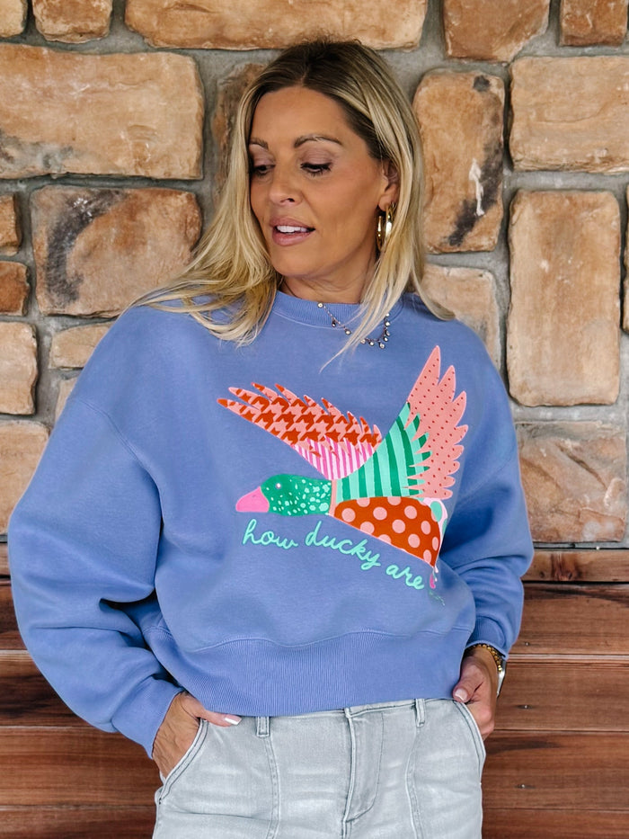 A woman wearing a light blue crewneck sweatshirt with a colorful patchwork duck graphic and the text 'how ducky are you' on the front, paired with light gray jeans, standing in front of a rustic stone wall.