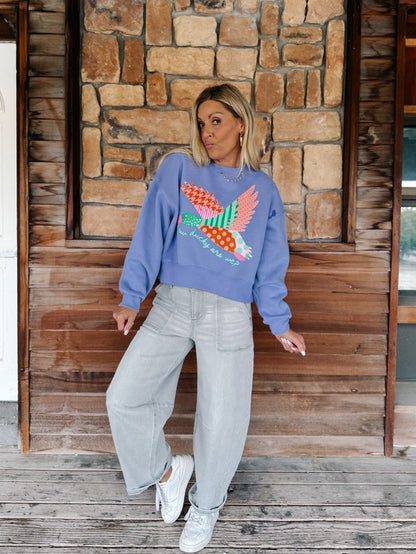 A woman stands on a rustic wooden porch wearing a light blue crewneck sweatshirt with a colorful patchwork bird design, paired with light gray wide-leg jeans and white sneakers.