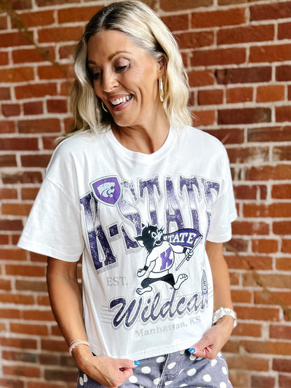 A woman wearing a white graphic t-shirt with purple and black 'K-STATE Wildcats' text and a wildcat mascot, standing in front of a red brick wall.