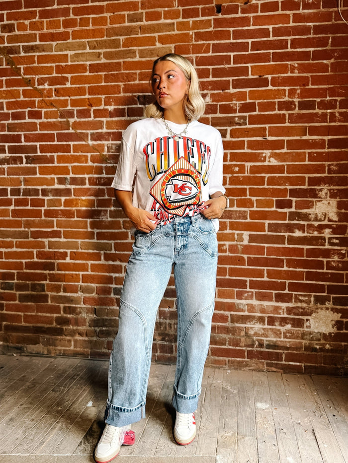 A woman stands against a red brick wall wearing a white graphic t-shirt with a colorful 'Chile' band print, light-wash blue jeans, and white sneakers.