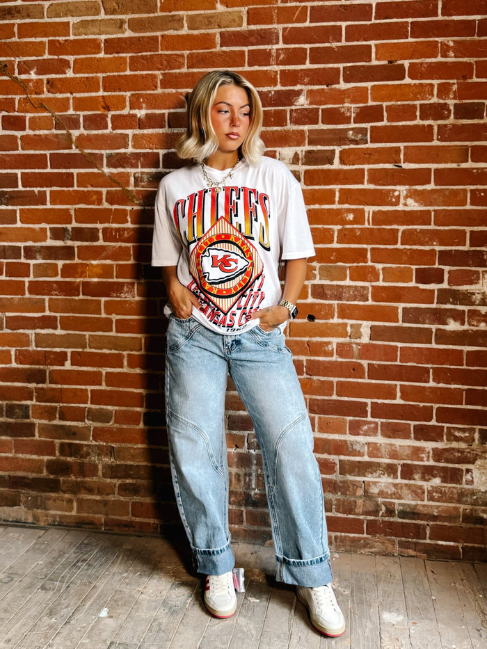 A woman stands against a red brick wall wearing a white Kansas City Chiefs graphic t-shirt, light-wash wide-leg jeans, and white sneakers.