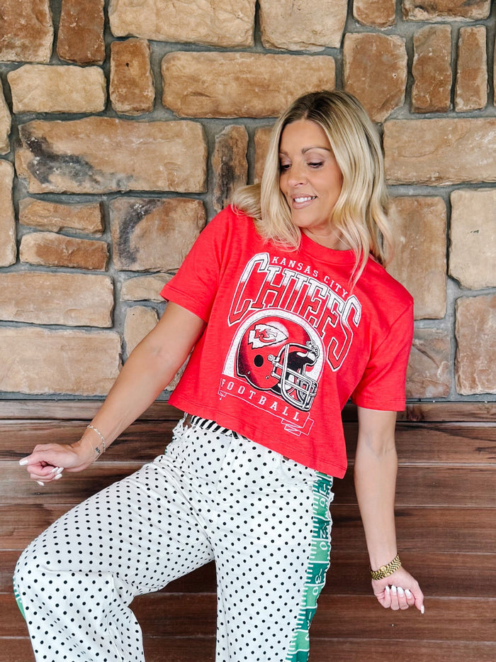 A woman wearing a red Kansas City Chiefs graphic t-shirt and white polka dot pants with green fringe, posing in front of a rustic stone wall.