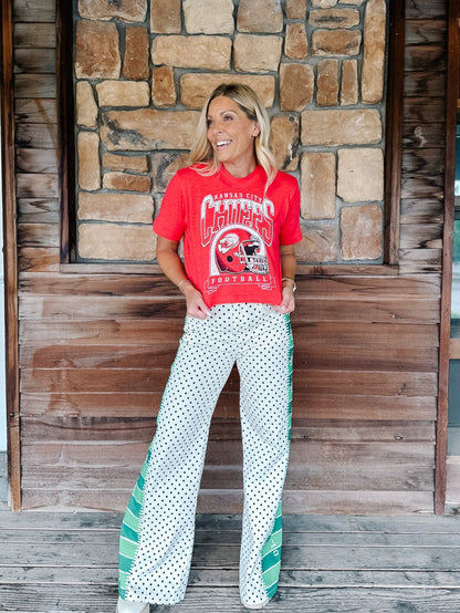 A woman stands on a wooden porch in front of a stone wall, wearing a red 'Kansas State Cowboys Football' graphic t-shirt and white wide-leg pants with green and white polka dots and side stripes.