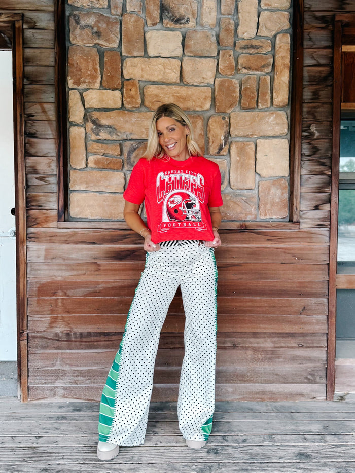 A woman stands on a wooden porch in front of a stone wall, wearing a red graphic t-shirt and white wide-leg pants with green and black polka dots and green side panels.