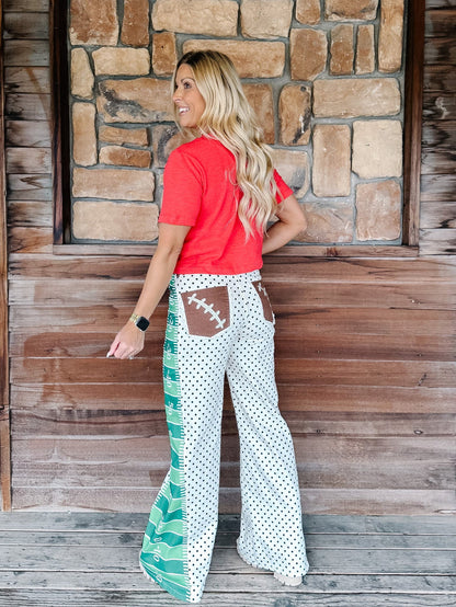 A woman stands on a wooden porch in front of a stone wall, wearing wide-leg white pants with black polka dots, brown football-shaped back pockets, and green and white side panels, paired with a coral red t-shirt.
