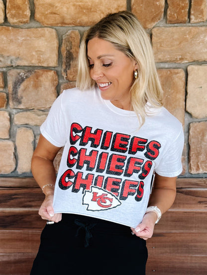 A woman wearing a white graphic t-shirt with 'CHIEFS CHIEFS CHIEFS' printed in red and black, and the Kansas City Chiefs logo below, standing in front of a stone wall.