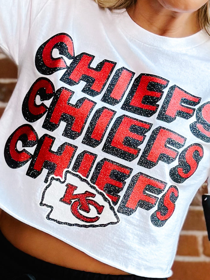 A close-up of a person wearing a white cropped T-shirt with large red and black 'CHIEFS' lettering and a Kansas City Chiefs logo graphic on the front.
