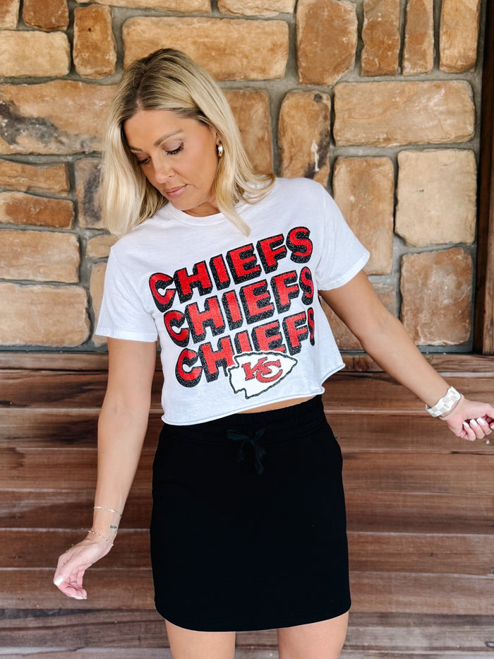 A woman wearing a white Kansas City Chiefs cropped graphic t-shirt with red and black lettering, paired with a black mini skirt, standing in front of a rustic stone wall.