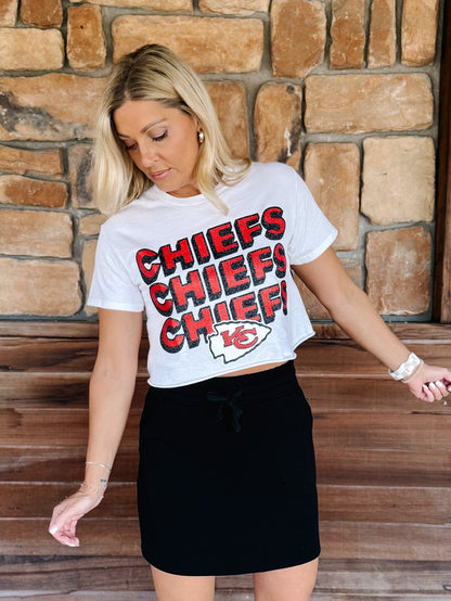 A woman wearing a white Kansas City Chiefs cropped graphic t-shirt with red and black lettering, paired with a black mini skirt, standing in front of a rustic stone wall.