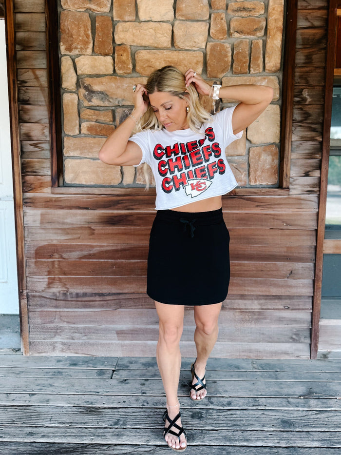 A woman stands on a rustic wooden porch wearing a white cropped graphic T-shirt with 'CHIEFS CHIEFS KC' in red and black lettering, paired with a black mini skirt and black strappy sandals.