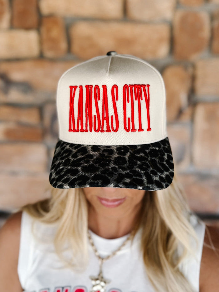 A woman wearing a snapback cap with a cream crown, red 'KANSAS CITY' text on the front, and a black leopard-print visor, posed against a blurred brick wall.