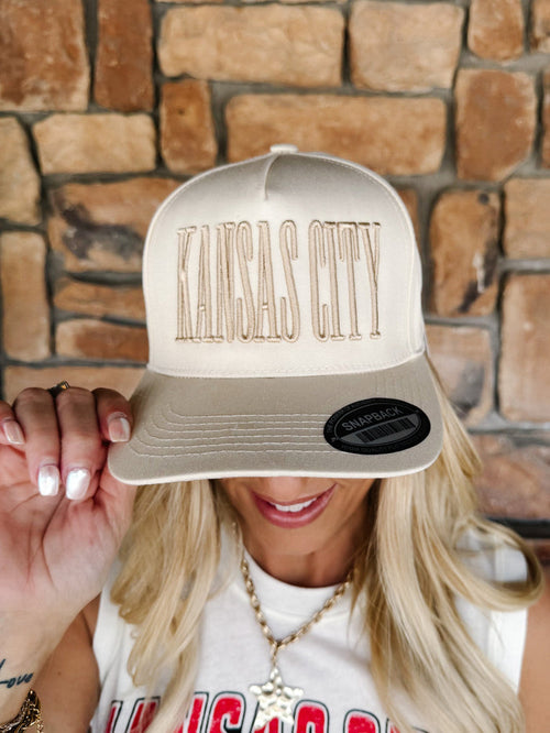A woman wearing a cream-colored snapback cap with 'KANSAS CITY' embroidered in a matching beige tone across the front. She is holding the brim of the hat with one hand, showing off the fit, against a rustic brown stone wall background.