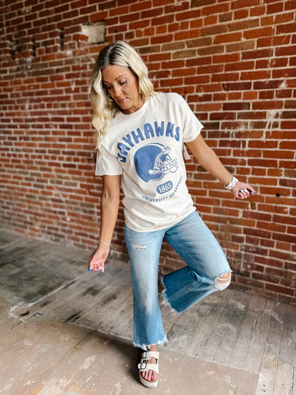 A woman posing in a white graphic t-shirt with a blue 'Jayhawks' football helmet design, paired with distressed blue jeans and white slide sandals, set against a red brick wall background.