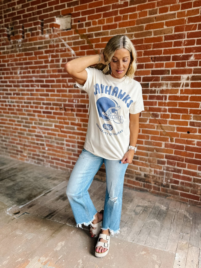 A woman posing in front of a brick wall wearing a cream-colored graphic t-shirt with a blue 'JAYHANKS' football helmet design, light-wash distressed jeans, and white slide sandals.