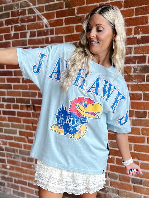 A woman wearing an oversized light blue graphic t-shirt with 'JAYHAWK' and the Kansas Jayhawks logo, posed against a red brick wall.