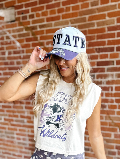 A woman wearing a white and purple 'K-STATE' baseball cap and a matching cream sleeveless graphic top with 'Wildcats' text, posing in front of a red brick wall.