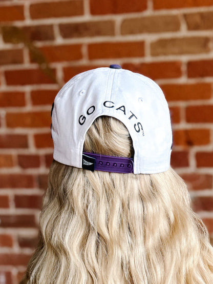 A white baseball cap with 'GO CATS' printed in purple on the back, worn by a person with long blonde hair in front of a red brick wall.