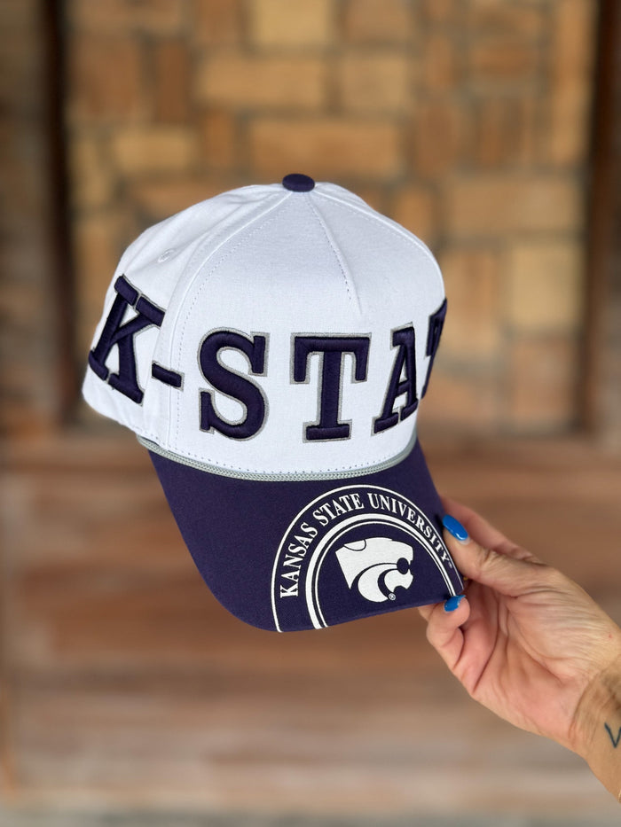 A white and purple blue baseball cap with 'K-STATE' embroidered on the front and a Kansas State University logo on the brim, held by a hand against a blurred brick background.