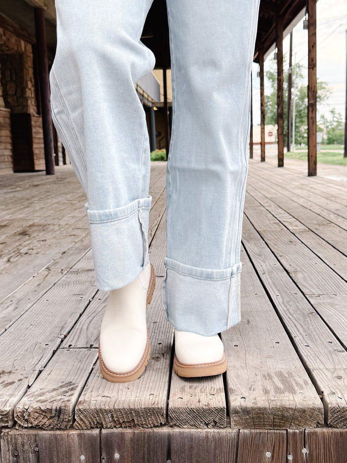 A person wearing light-wash blue jeans and off-white ankle boots with tan soles, standing on a weathered wooden boardwalk.