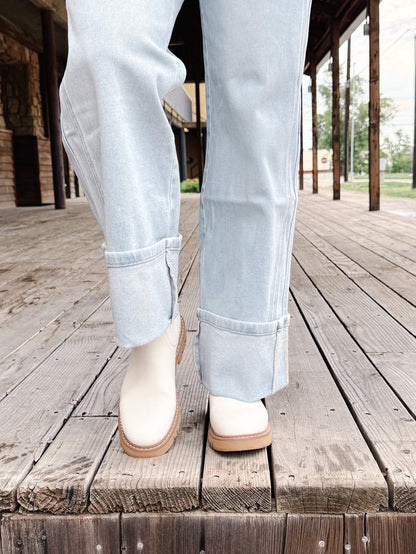 A person wearing light-wash blue jeans and off-white ankle boots with tan soles, standing on a weathered wooden boardwalk.