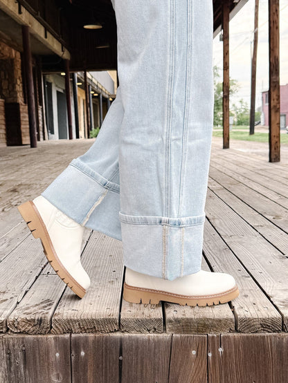 A person wearing light blue cuffed jeans and off-white ankle boots with tan soles, standing on a rustic wooden deck beside a porch.