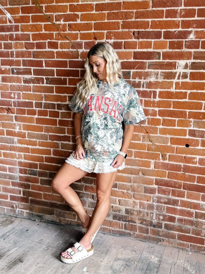 A woman posing against a red brick wall wearing a camouflage graphic t-shirt with 'Kansas' printed in red, paired with a white ruffled mini skirt and white slide sandals.