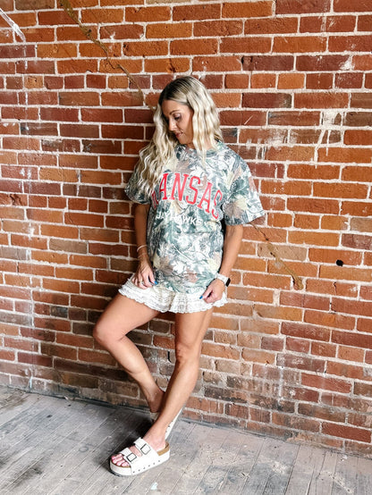 A woman posing against a red brick wall wearing a camouflage graphic t-shirt with 'Kansas' printed in red, paired with a white ruffled mini skirt and white slide sandals.