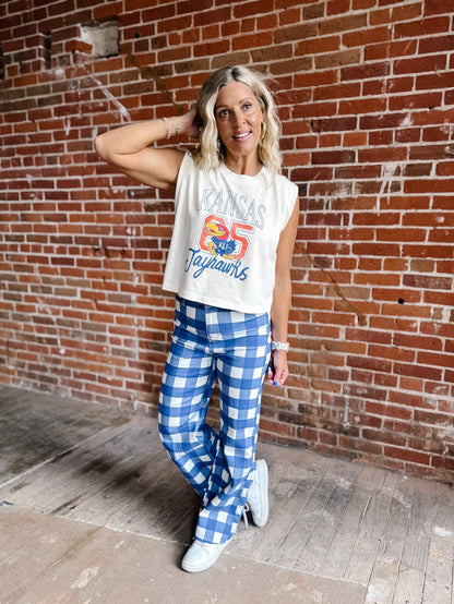 A woman posing in front of a red brick wall wearing a cream sleeveless crop top with a 'Kansas Jayhawks' graphic and blue-and-white gingham pants.