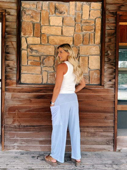 A woman stands on a rustic wooden porch in front of a stone wall, wearing light blue wide-leg cargo pants, a white tank top, and brown sandals.