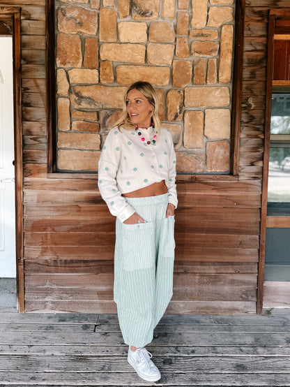 A woman stands on a rustic wooden porch in front of a stone wall, wearing a cream cropped sweater with mint green dots, light green pinstriped wide-leg pants, and white sneakers.