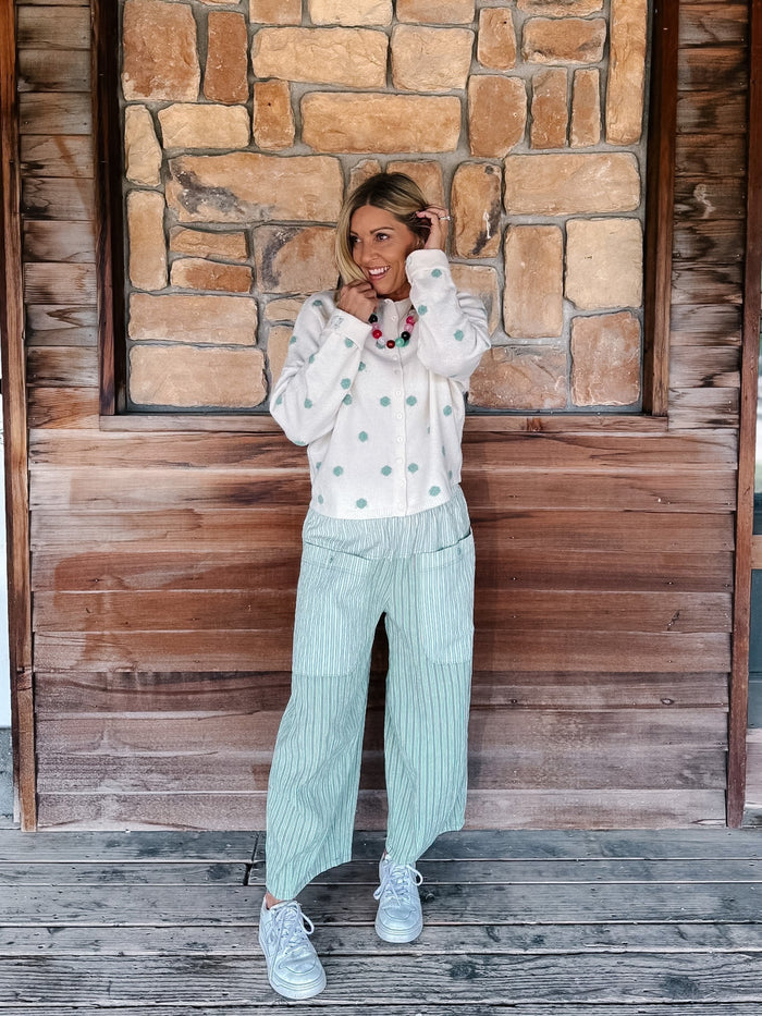 A woman stands on a wooden deck in front of a rustic stone and wood wall, wearing a white long-sleeve button-up shirt with mint green buttons, light green pinstriped wide-leg pants, and white sneakers.