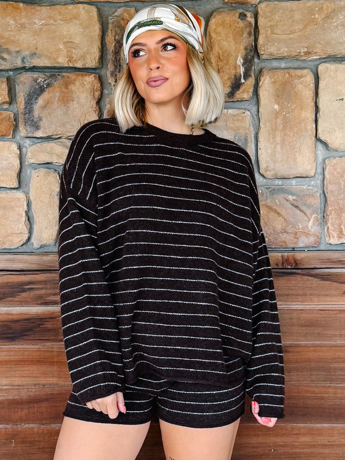 A woman wearing a brown knit long-sleeve top with thin white horizontal stripes and matching brown shorts, standing in front of a rustic stone wall and wooden bench.