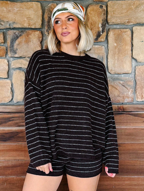 A woman wearing a brown knit long-sleeve top with thin white horizontal stripes and matching brown shorts, standing in front of a rustic stone wall and wooden bench.