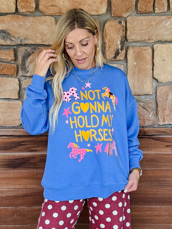 A woman wearing a bright blue crewneck sweatshirt with a colorful graphic that reads 'NOT GONNA HOLD MY HORSES' with pink and yellow horses and stars, paired with red and white polka dot pants, standing in front of a rustic stone wall.