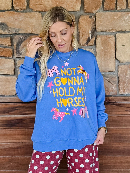 A woman wearing a bright blue crewneck sweatshirt with a colorful graphic that reads 'NOT GONNA HOLD MY HORSES' with pink and yellow horses and stars, paired with red and white polka dot pants, standing in front of a rustic stone wall.