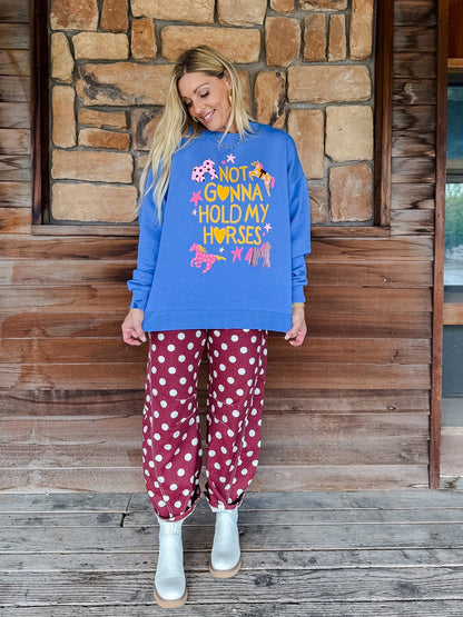 A woman stands on a wooden porch in front of a stone wall, wearing a bright blue sweatshirt with a colorful graphic that reads 'NOT GONNA HOLD MY HORSES' and matching red pants with white polka dots.