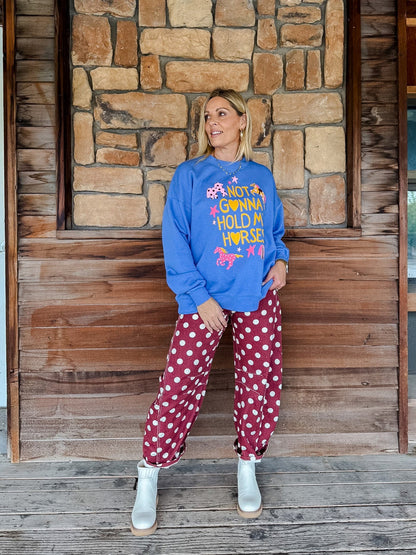 A woman stands on a wooden porch in front of a stone wall, wearing a bright blue sweatshirt with a colorful graphic and red-and-white polka dot pants.
