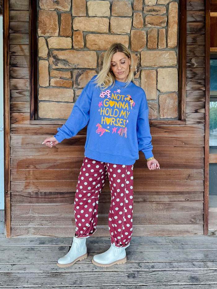A woman stands on a wooden porch in front of a stone wall, wearing a bright blue sweatshirt with a colorful graphic that reads 'I LOVE YOU HOLD MY HORSY!' and red pants with white polka dots.