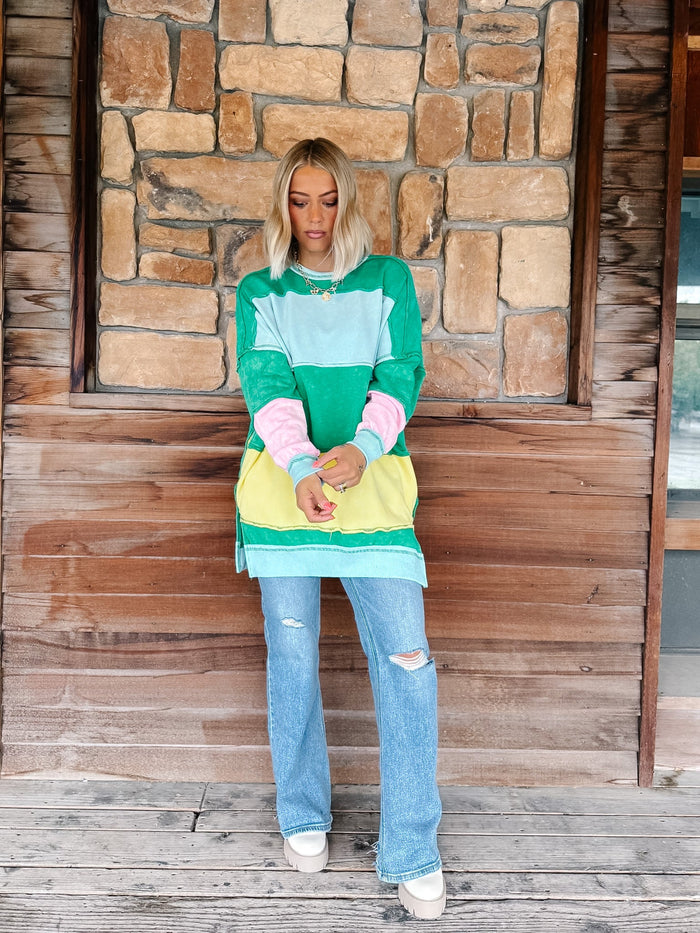 A woman stands on a rustic wooden porch in front of a stone wall, wearing a colorful color-blocked sweatshirt with green, light blue, pink, and yellow panels, paired with light-wash distressed jeans and white sneakers.