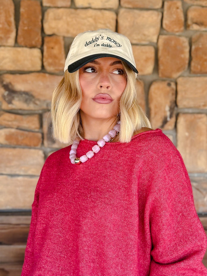 A woman with blonde bob-cut hair wearing a cream baseball cap with black embroidery that reads 'Daddy's money is mine', a bright pink knit sweater, and a pink beaded necklace, standing in front of a rustic stone wall.